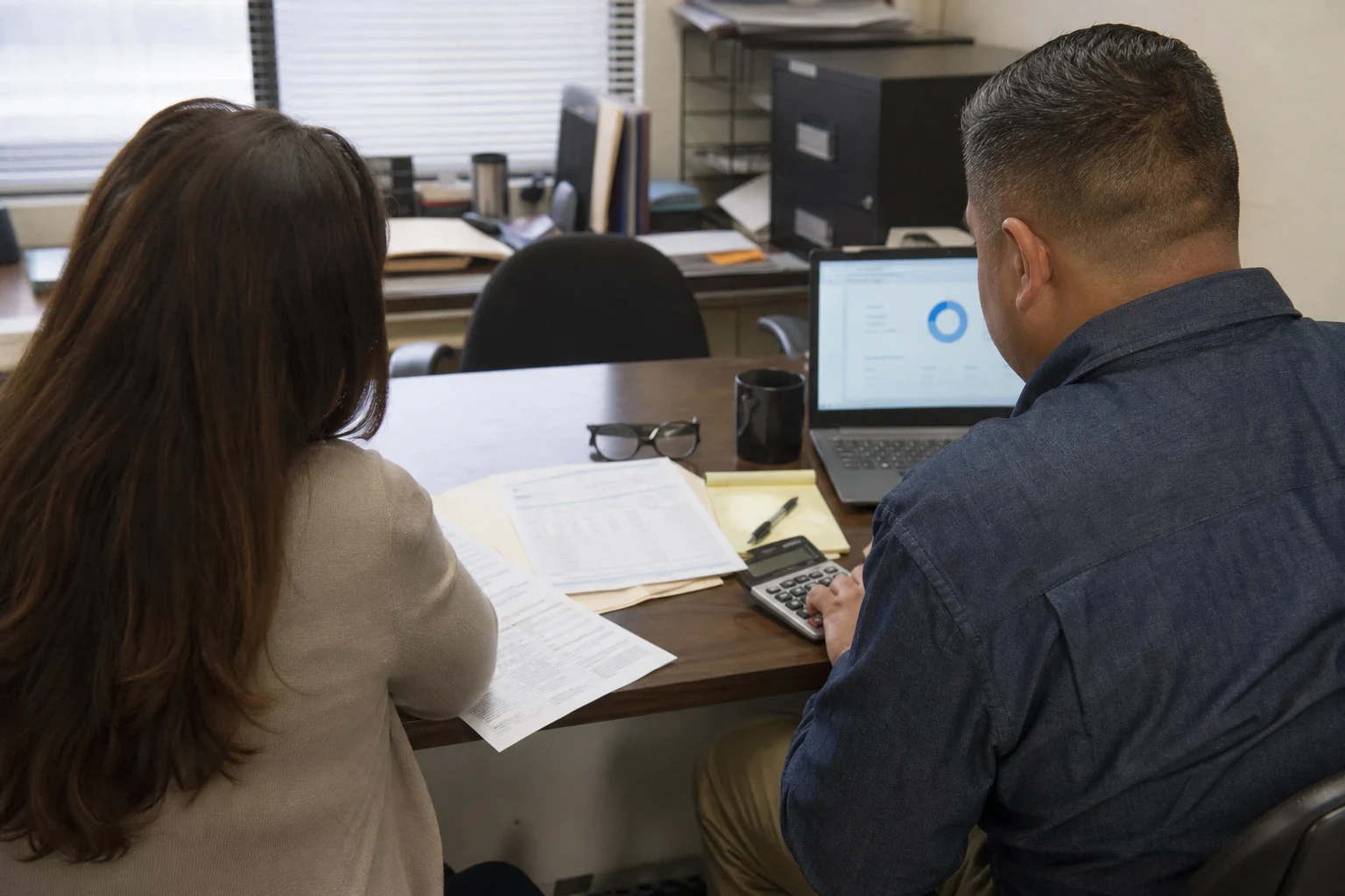 Two people seen from behind at an office desk reviewing printed statements, one tapping a calculator beside a notepad, laptop and coffee mug, with a filing cabinet nearby.