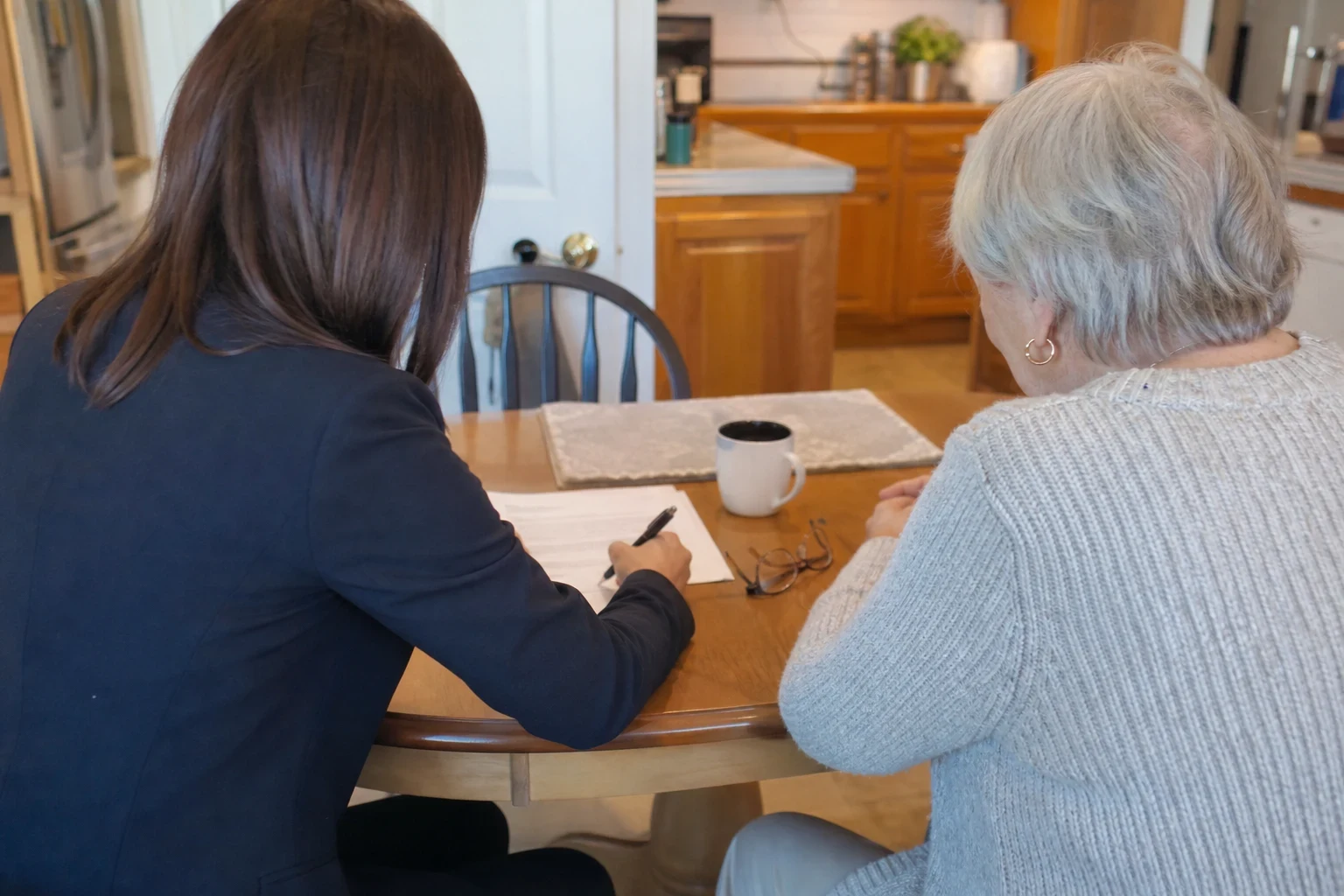 An agent in a navy blazer writes on a paper form at a round wooden kitchen table beside a gray-haired woman, with reading glasses and a coffee mug on the table
