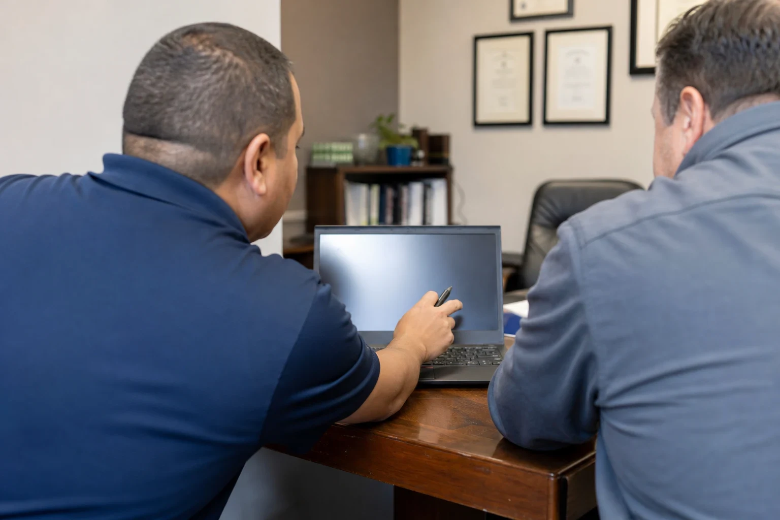 Two men seen from behind at a wooden office desk, one pointing a pen at a laptop screen, with framed certificates, a bookshelf and a leather chair behind