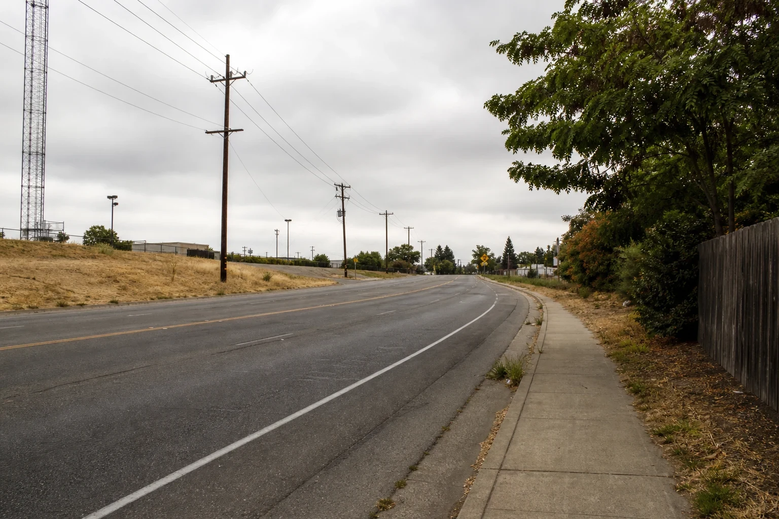 Empty curving road on the edge of town under an overcast sky, with utility poles, dry grass, a lattice tower, a sidewalk and a wooden fence lined with trees